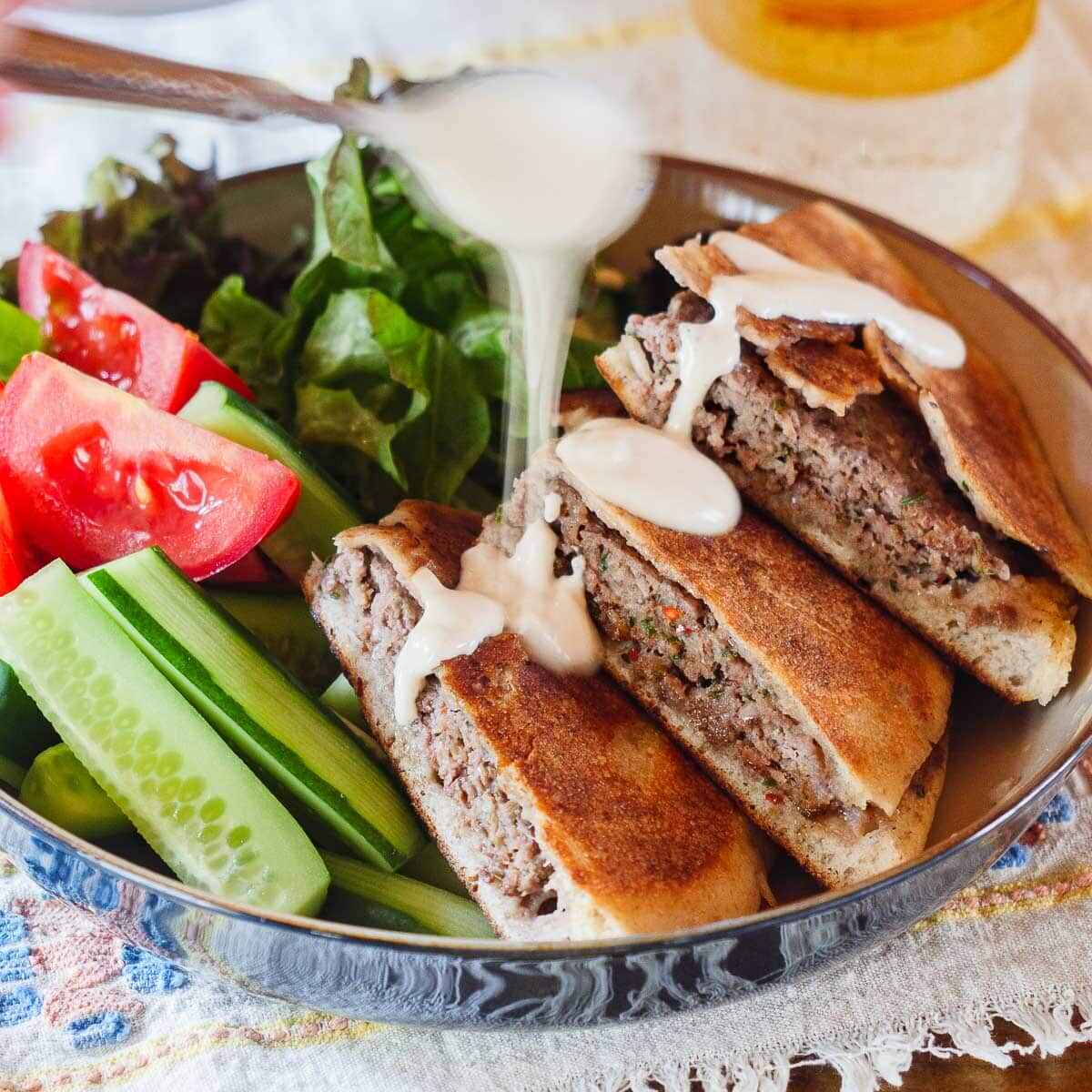 سس تاهینی را روی آرایه های طلایی در یک کاسه با گوجه فرنگی تازه خرد شده، خیار و جعفری تازه بریزید. تصویر از منظر سطح چشم دیده می شود.