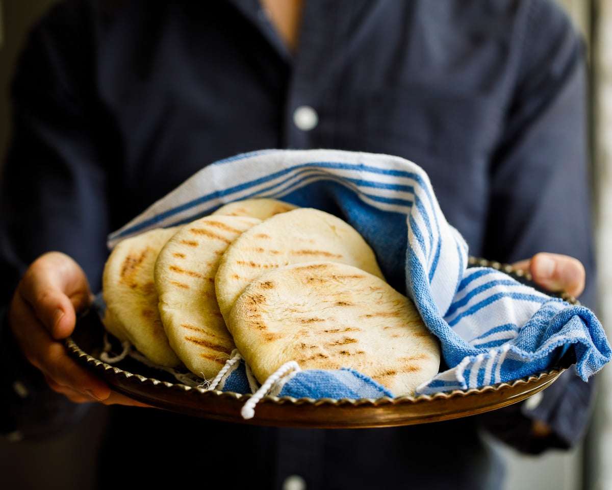 نان پیتا روی یک سینی نگه داشته شده خرید یک نان اصلی با یک پیراهن تیره. زاویه تقریباً افقی به نان پیتا است که در پارچه آبی و سفید ترکی پیچیده شده است.